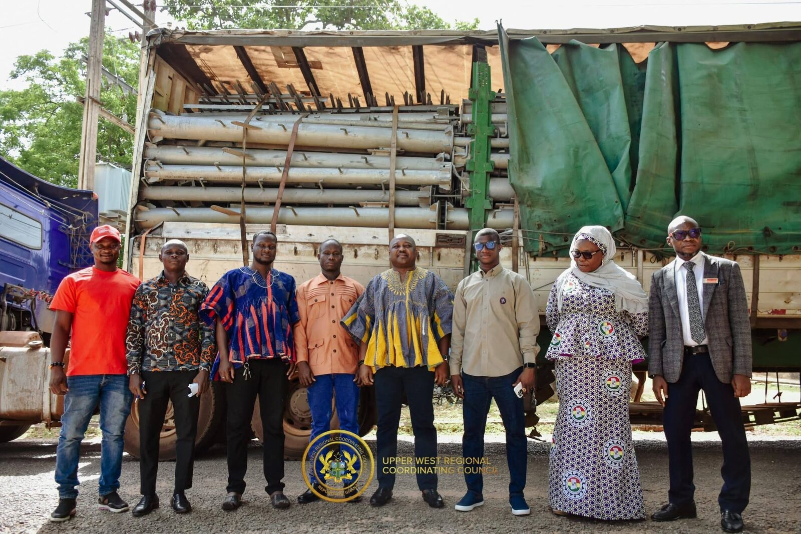 A group of eight people standing in a row in front of a lorry loaded with long metal pipes, wearing a mix of colourful clothes.