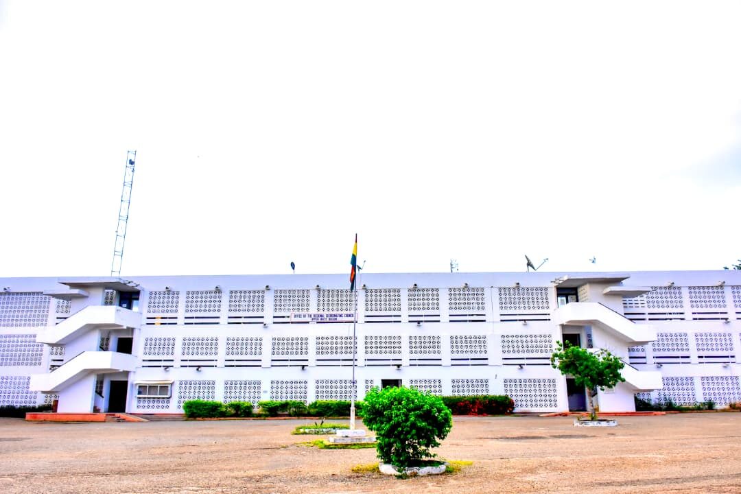 Front view of the white multi-storey office building with patterned walls, a flag at the centre and shrubs in the open yard.